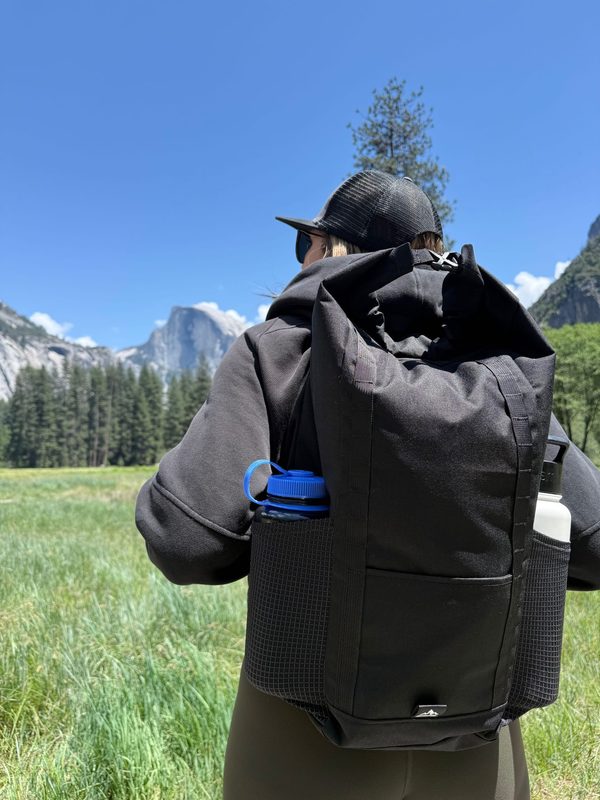Rear view of the black 17L Rolltop Daypack being worn in Yosemite Valley, with bottles in both exterior side pockets
